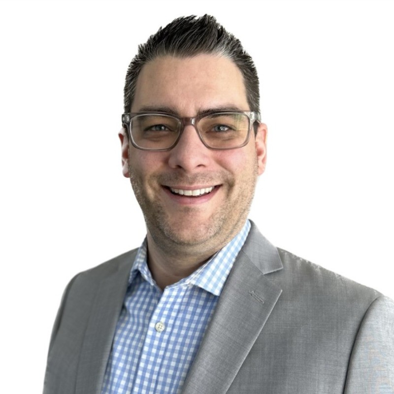 A man with short dark hair and glasses, wearing a light gray suit jacket over a blue and white checkered shirt, smiles in front of a plain white background.