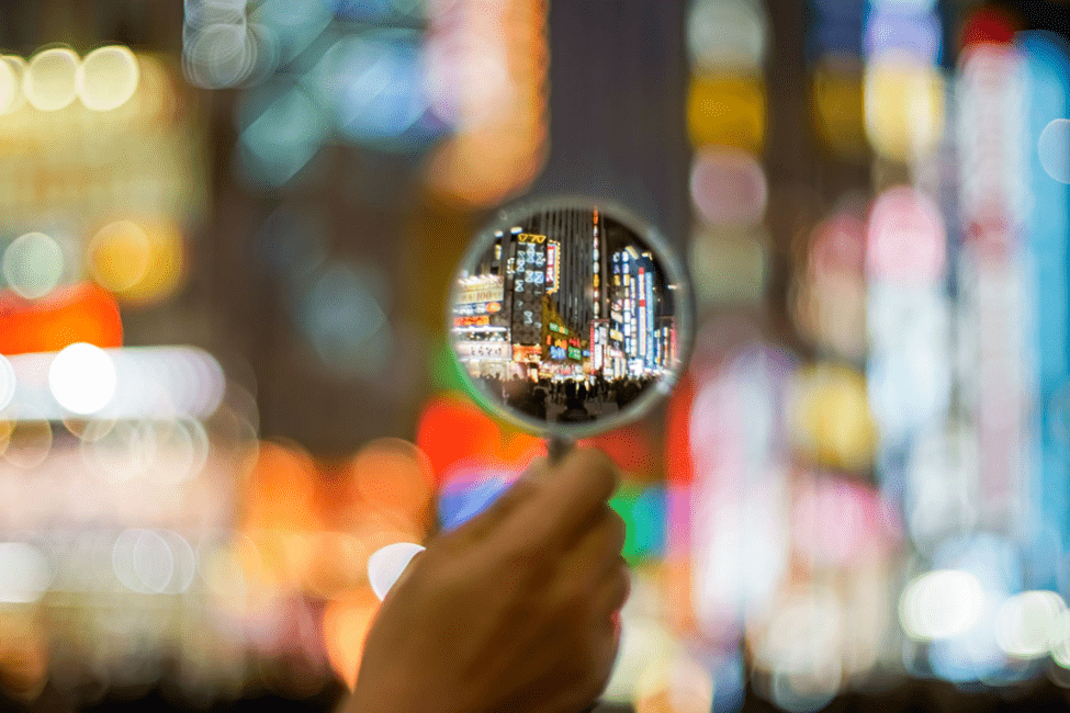 A hand holds a magnifying glass that focuses on a busy, brightly lit urban street filled with neon signs and colorful lights. The background outside the magnified area is blurred, highlighting the cityscape viewed through the magnifying glass.