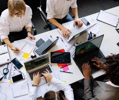 Four people work at a cluttered table with laptops, notebooks, papers, smartphones, pens, and a tablet, collaborating on a project in an office setting.