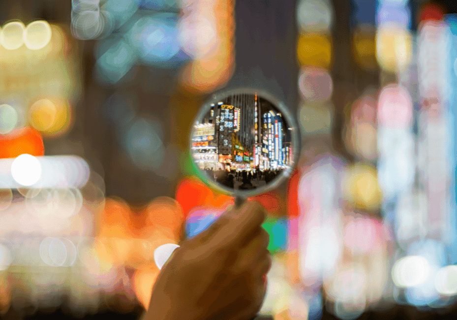 A hand holds a magnifying glass that focuses on a busy, brightly lit urban street filled with neon signs and colorful lights. The background outside the magnified area is blurred, highlighting the cityscape viewed through the magnifying glass.