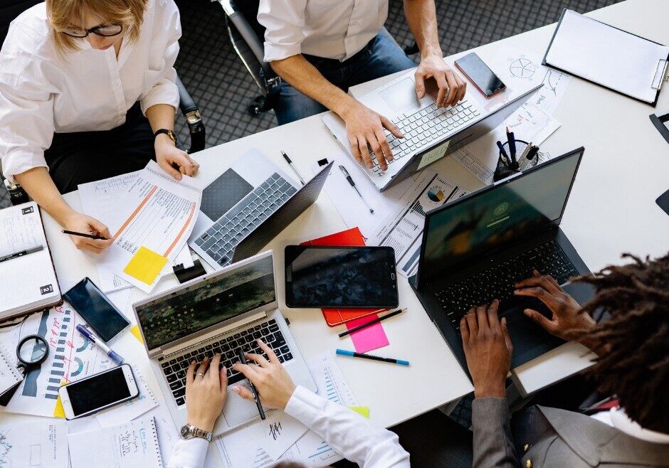 Four people work at a cluttered table with laptops, notebooks, papers, smartphones, pens, and a tablet, collaborating on a project in an office setting.