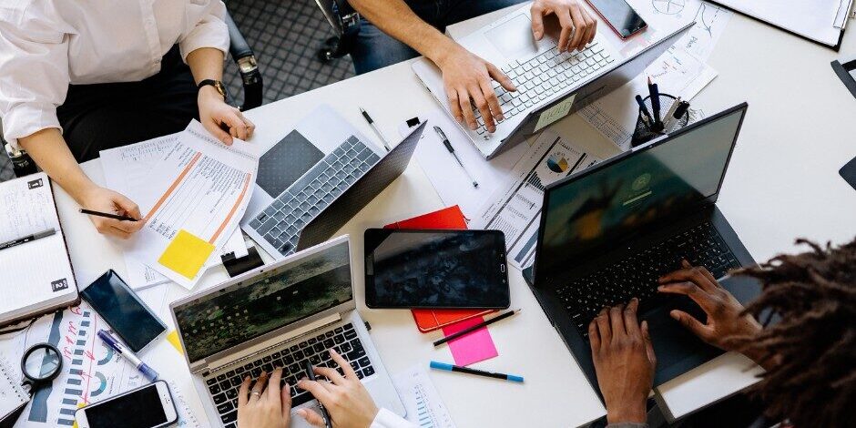 Precision Writing Four people work at a cluttered table with laptops, notebooks, papers, smartphones, pens, and a tablet, collaborating on a project in an office setting.