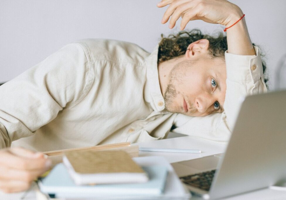 A person with long curly hair and a neutral expression is resting their head on their arm at a desk. They are wearing a light-colored shirt and are surrounded by notebooks, a pen, and a laptop. The scene suggests feeling tired or overwhelmed.