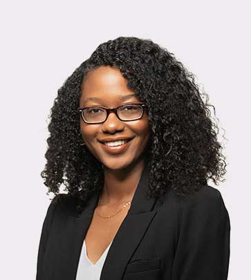 A woman with curly black hair and glasses smiles while wearing a black blazer over a light-colored top. She stands in front of a plain, light background.